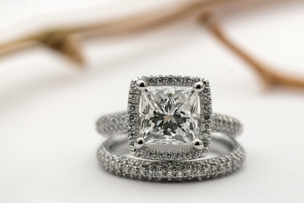 A close-up of a diamond engagement ring with a princess-cut center stone and a smaller diamond-studded band against a soft, blurred background.
