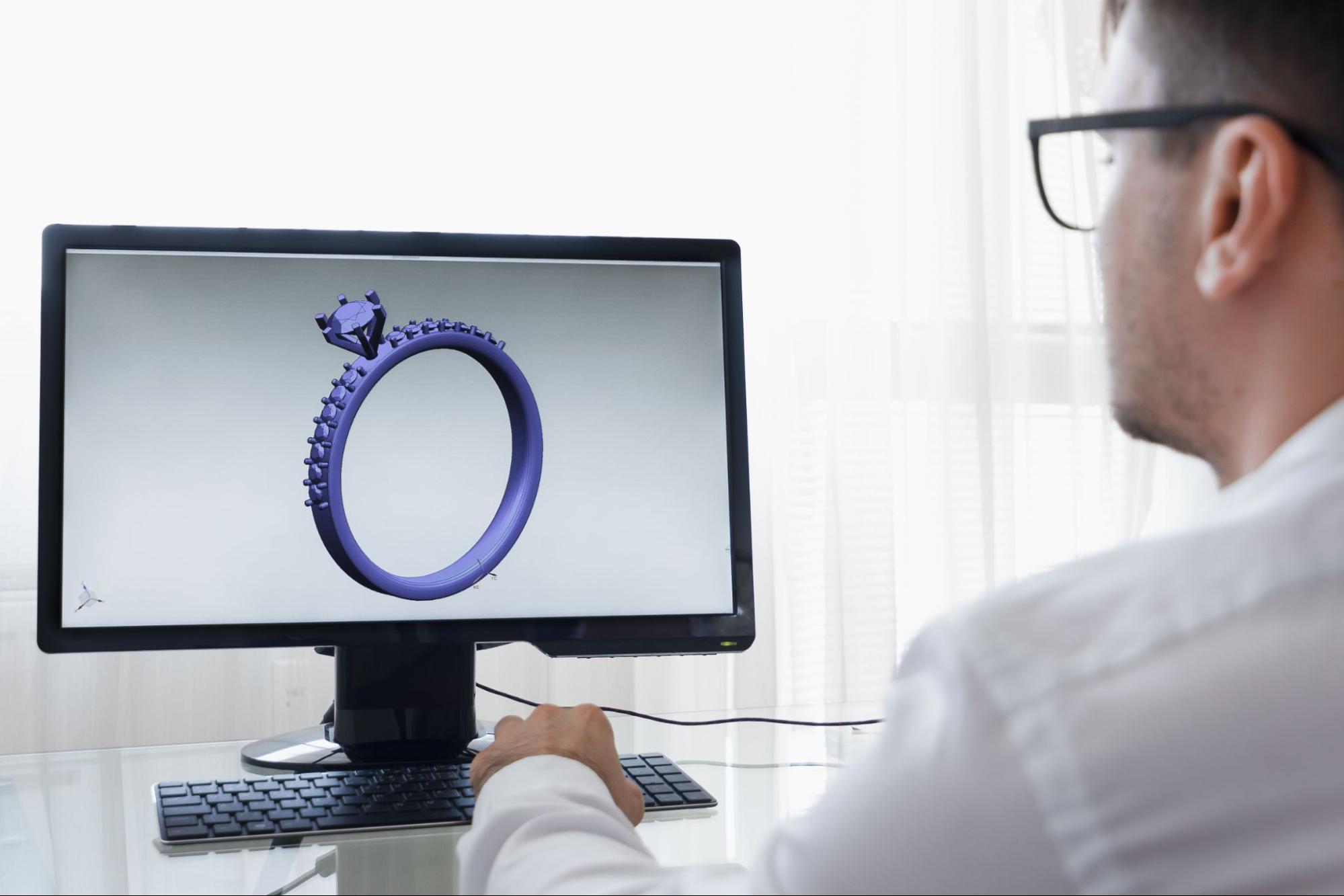 A jeweler in glasses views a detailed digital 3D model of a blue engagement ring on a computer screen.