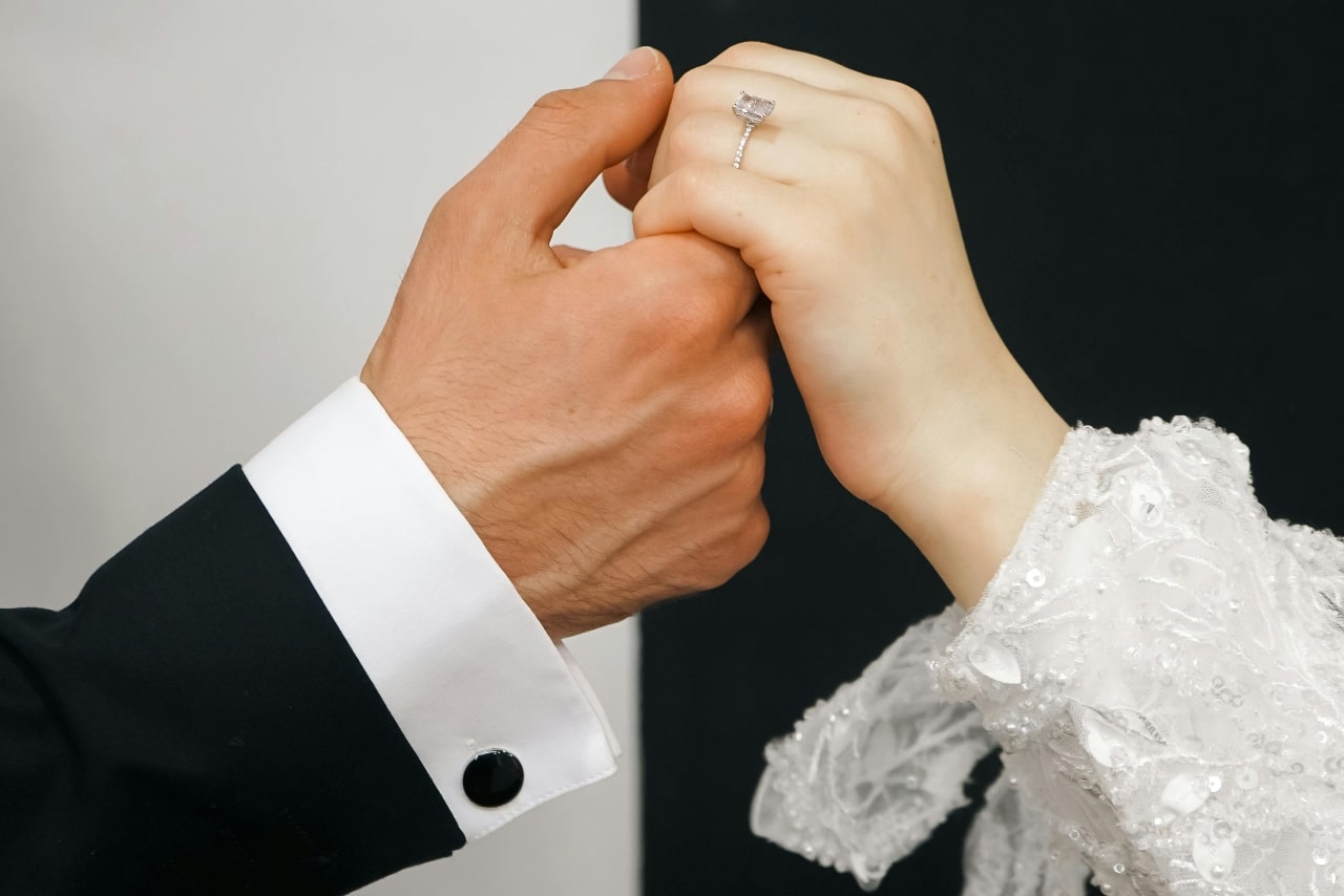 A groom in a black suit holds a bride's hand adorned with an engagement ring against a half black, half white background.