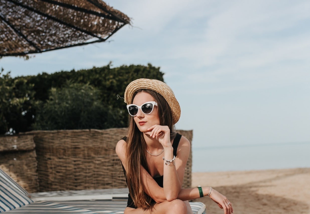 A woman wearing a straw hat, charm bracelet, chain necklace, and white sunglasses sits on a striped beach lounger by the seaside.