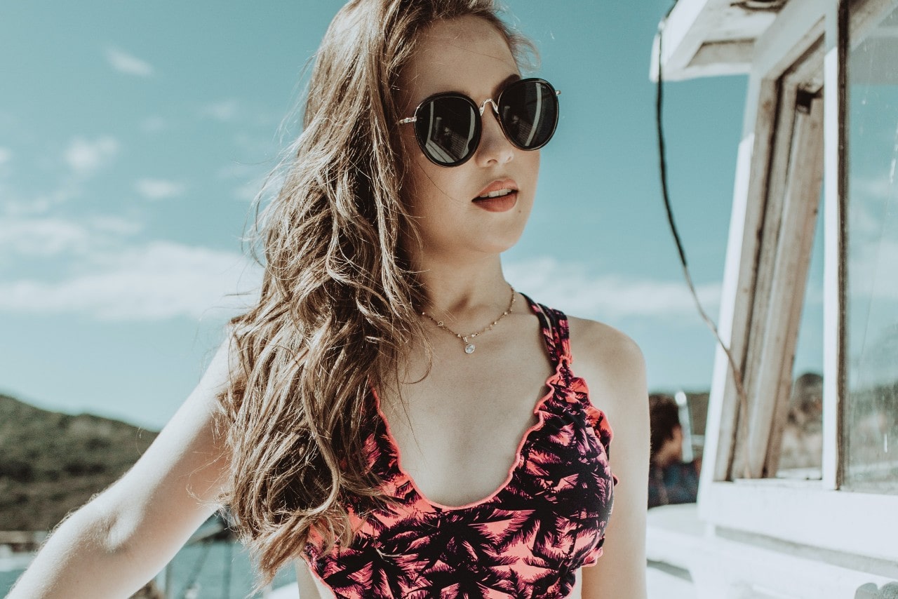 A woman in a pink printed swimsuit with black round sunglasses and yellow gold chain with a pearl stands against sunlight on a boat, surrounded by sea.