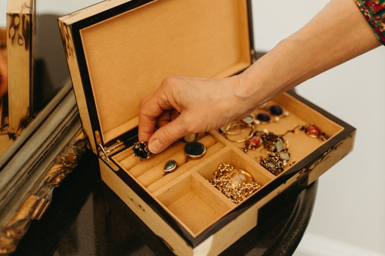 A view of a woman’s hand reaching into an open jewelry box filled with beautiful rings, bracelets, and earrings adorned with colorful gemstones.