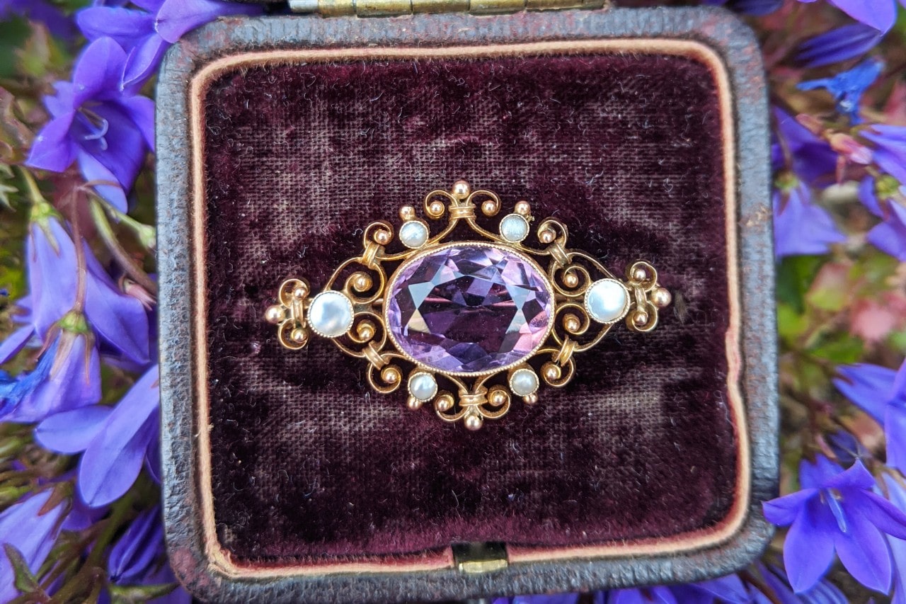 A close-up of an antique brooch with a large amethyst center stone and pearl accents set in intricate gold filigree on burgundy velvet, surrounded by vibrant purple flowers.