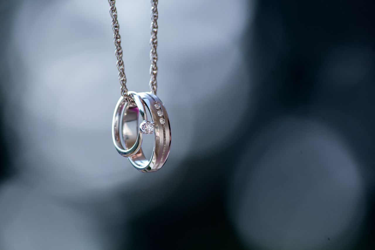 A white gold solitaire diamond engagement ring and a wedding band, hanging on a delicate chain, are set against a soft, blurred background.