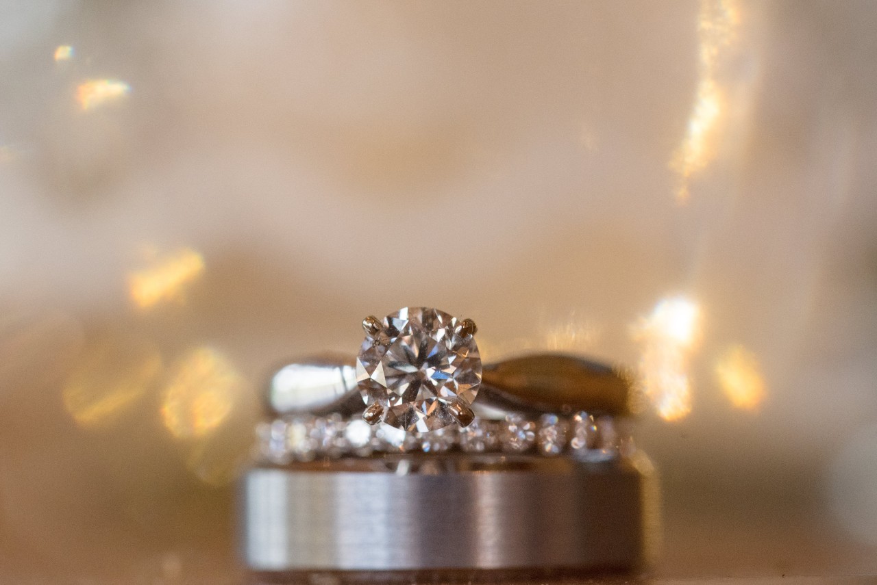 Close-up of a round cut diamond ring atop wedding bands, set against a warm, blurred background with soft golden lights.