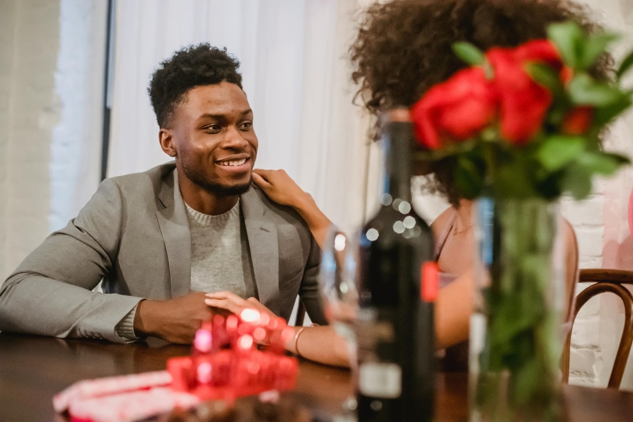 A smiling couple enjoys a romantic dinner together at a restaurant table decorated with red roses, wine, and wrapped gifts.