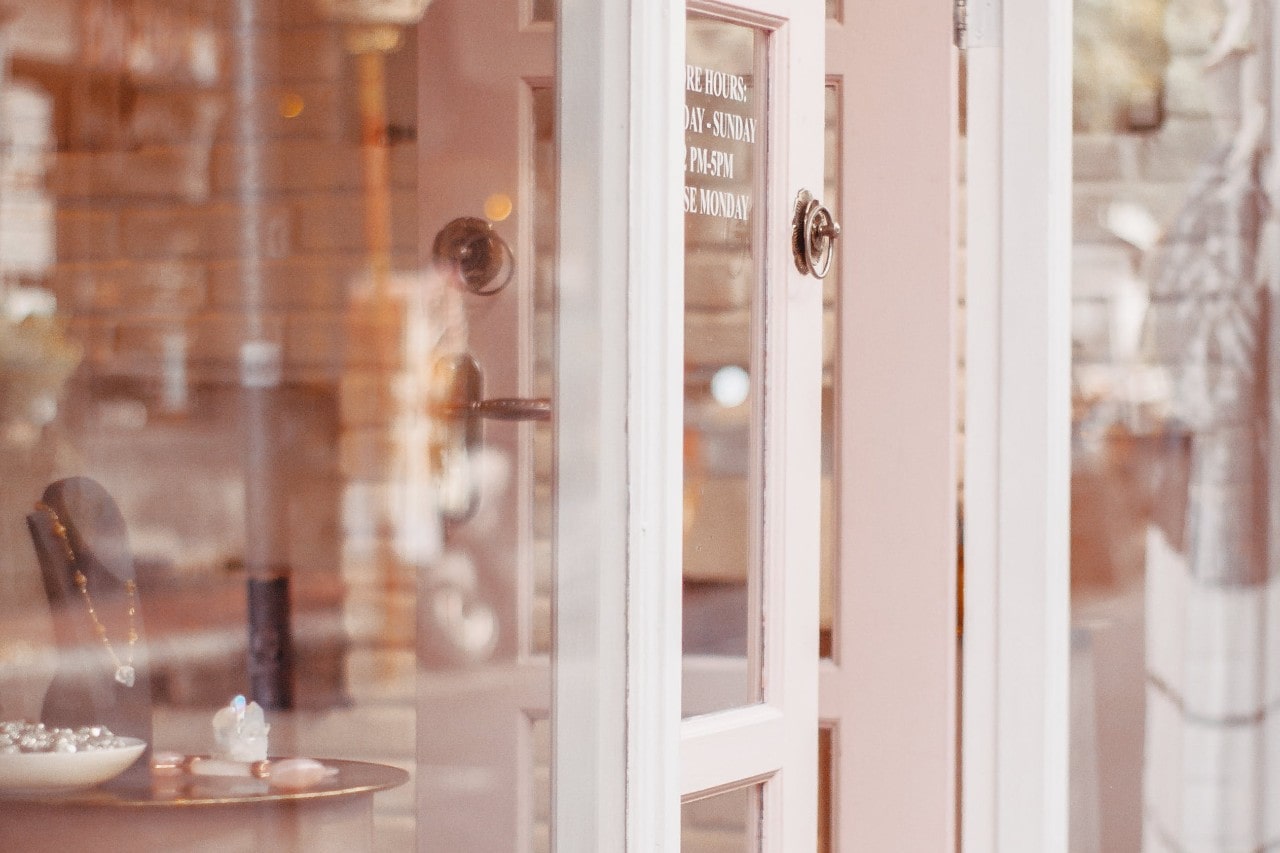 A soft-focus view of a boutique entrance with a partially open pink door and jewelry displayed inside.