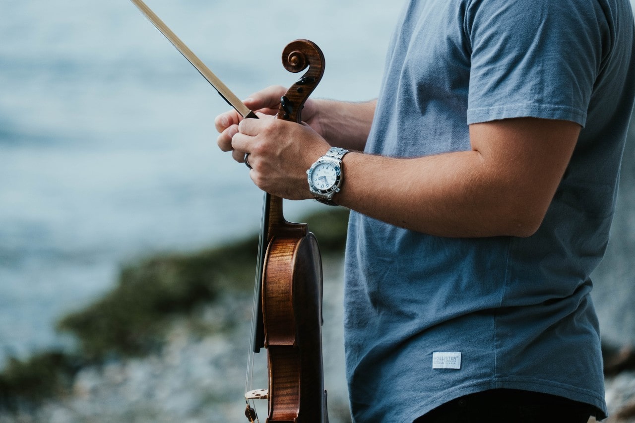 A close-up of a man wearing a stainless steel watch and holding a violin on a rocky shore.