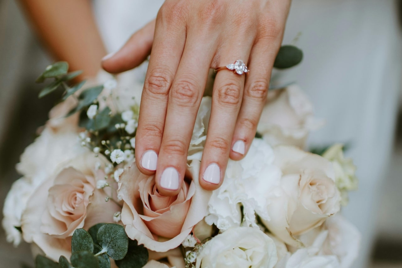 A close-up of a woman’s hand showing a three-stone engagement ring while resting on a bouquet of roses and greenery.