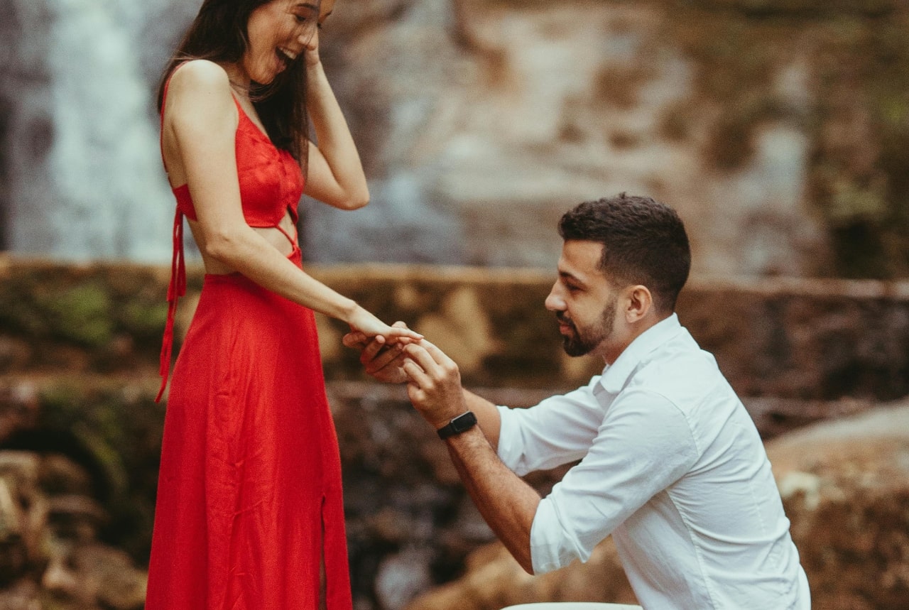A man kneels and places a ring on a smiling woman’s hand during an outdoor proposal by a waterfall.