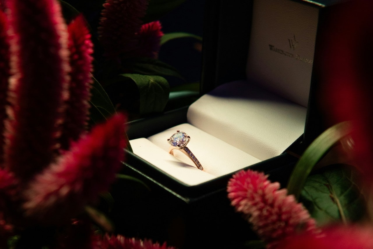 A rose gold diamond engagement ring displayed inside an open green velvet box surrounded by red flowers.