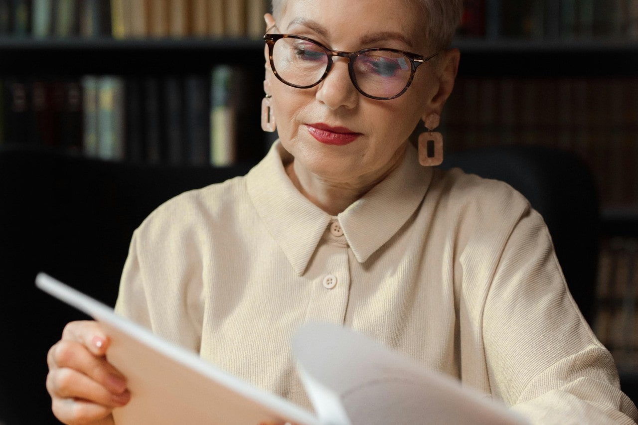 An elegant older woman with glasses and statement earrings reads a document in a library setting.