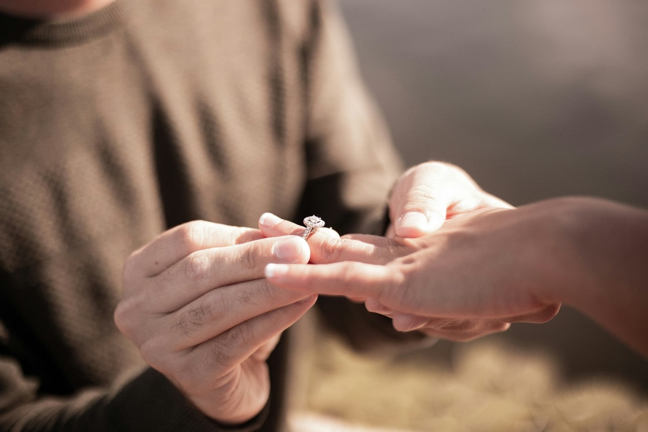 A man in brown weather puts a cushion cut diamond engagement ring on a woman’s finger in the outdoor sunny setting