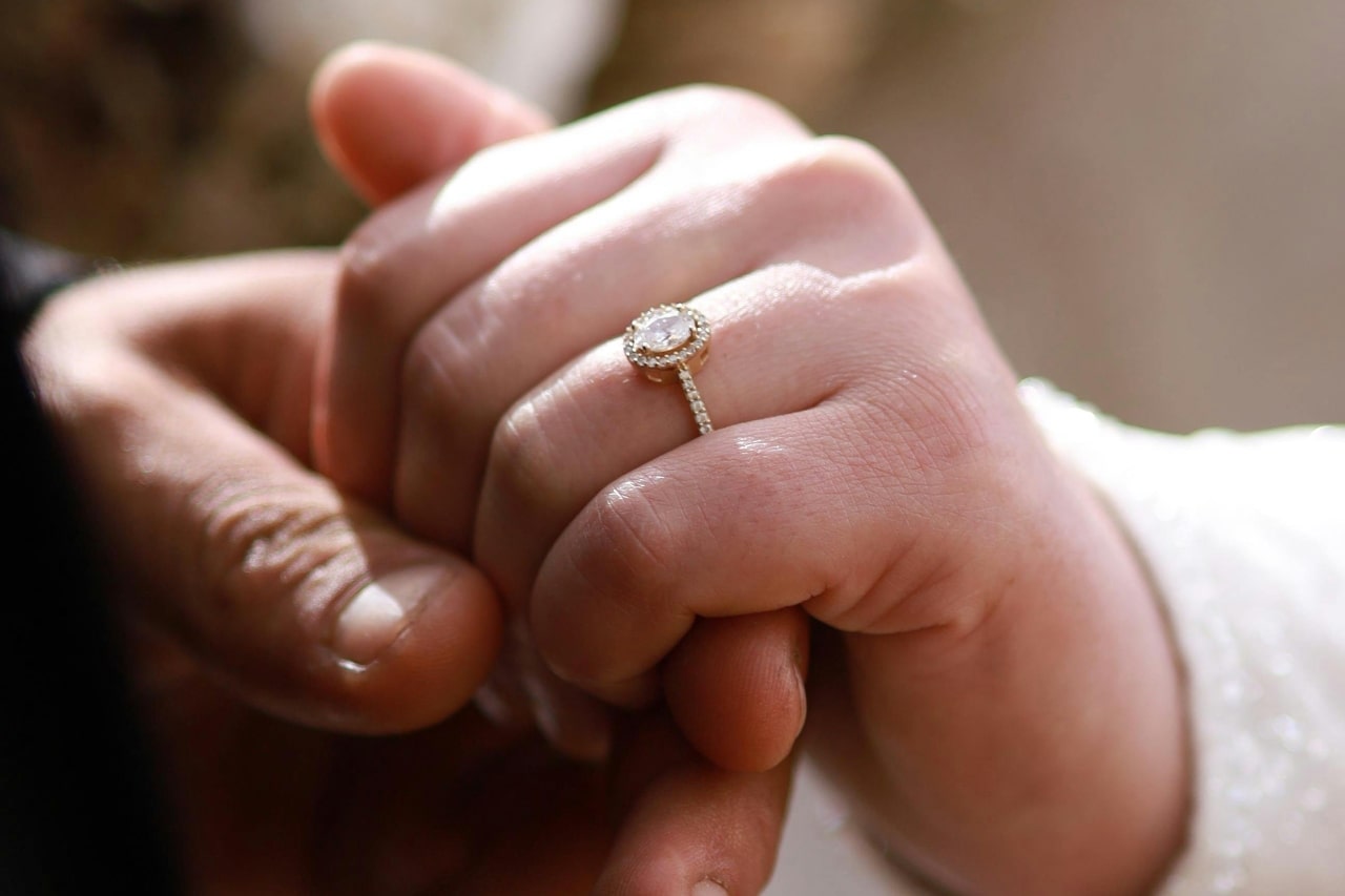 A close-up of a couple holding hands, the bride's hand wearing a white gold engagement ring featuring an oval halo setting.