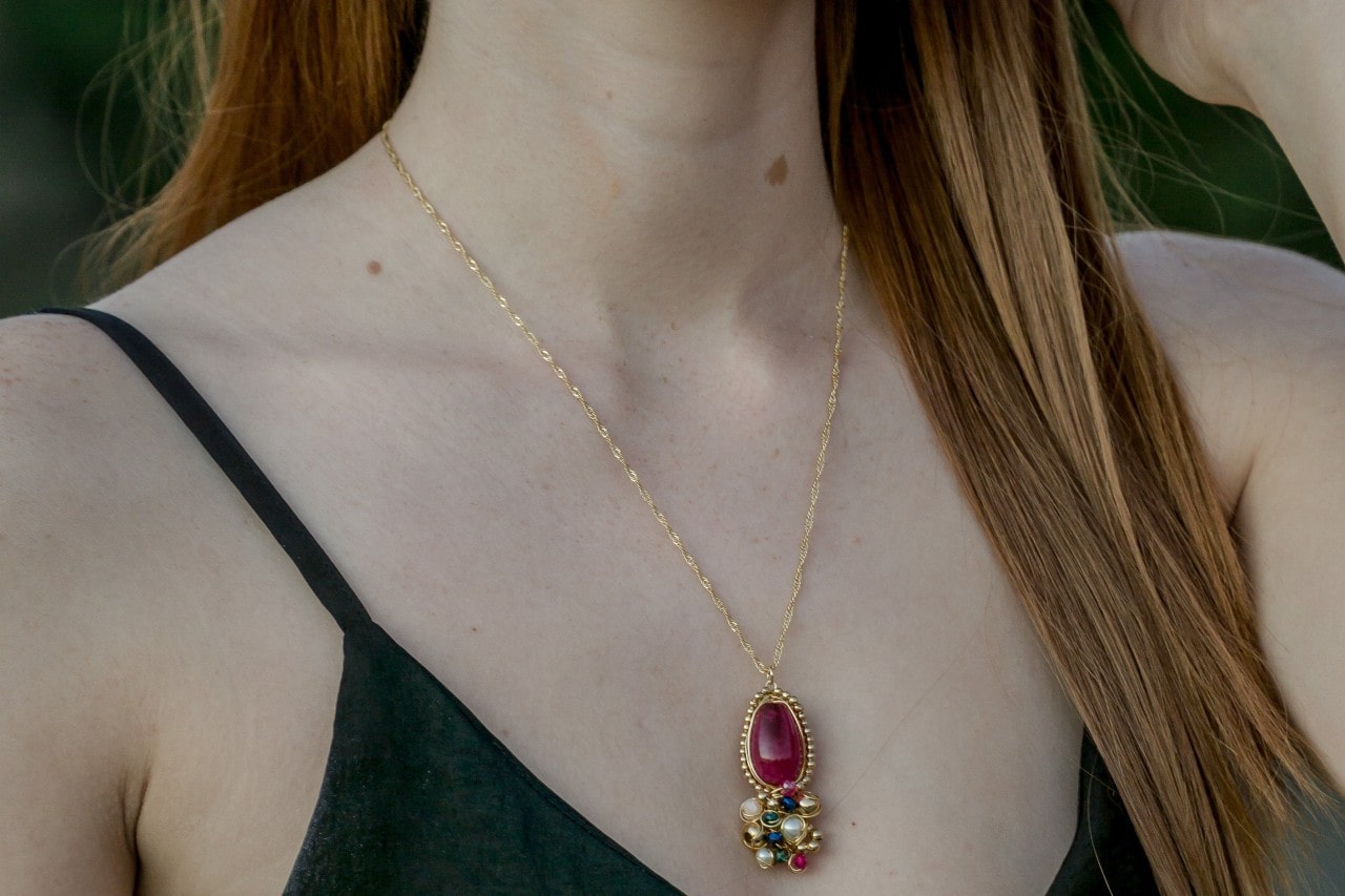 A woman with red hair wearing a long golden necklace with a large pendant made up of red, green, and pearl jewels.