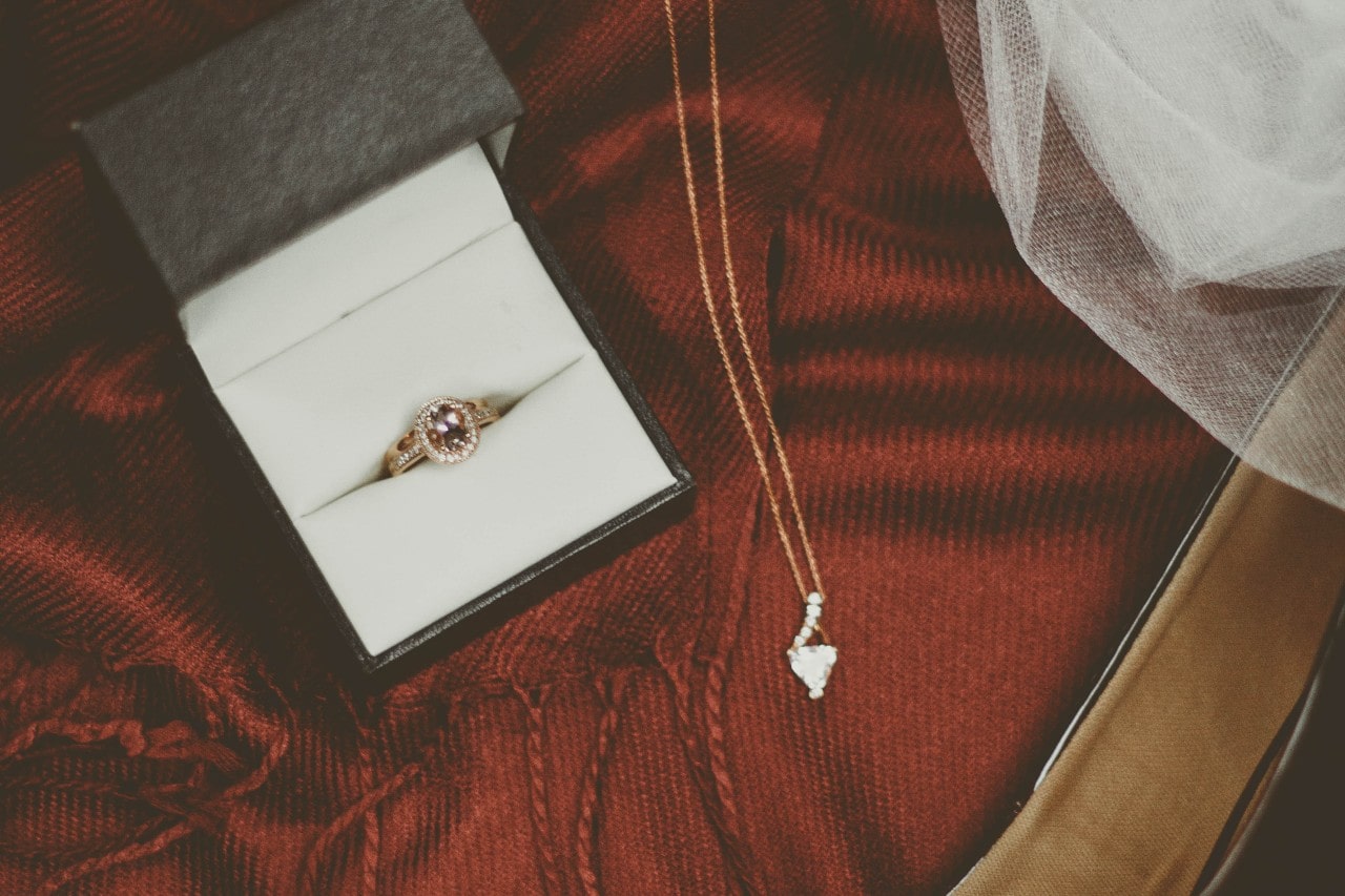A black ring box open with a golden ruby ring inside and a golden necklace lying next to it, both resting on red fabric.