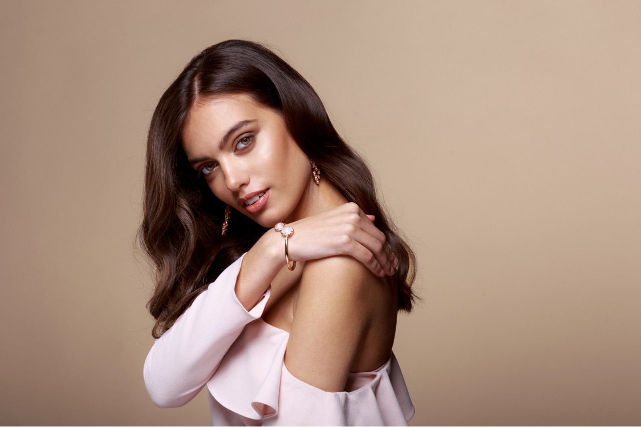 A woman with short brown hair in a shoulder-less pink top, featuring a rose gold bangle and dangle earrings.