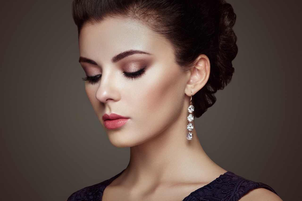 A woman with gorgeous make up and dangle diamond earrings against a dark background.