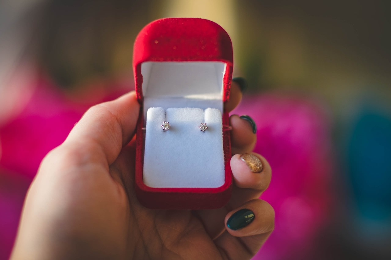A close-up of a hand holding a red velvet earring box with diamond studs inside.