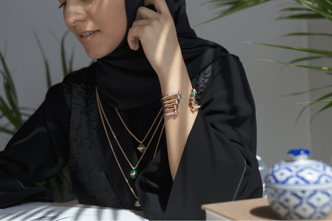 A woman looking down at a desk and resting her hand on the side of her head, showing off her rose gold bracelet and stacked rose gold necklaces.