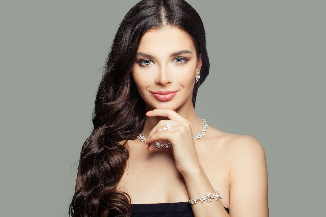 A woman with long dark hair poses against a neutral background, wearing a floral diamond necklace, matching earrings, bracelet, and rings, with her hand resting under her chin