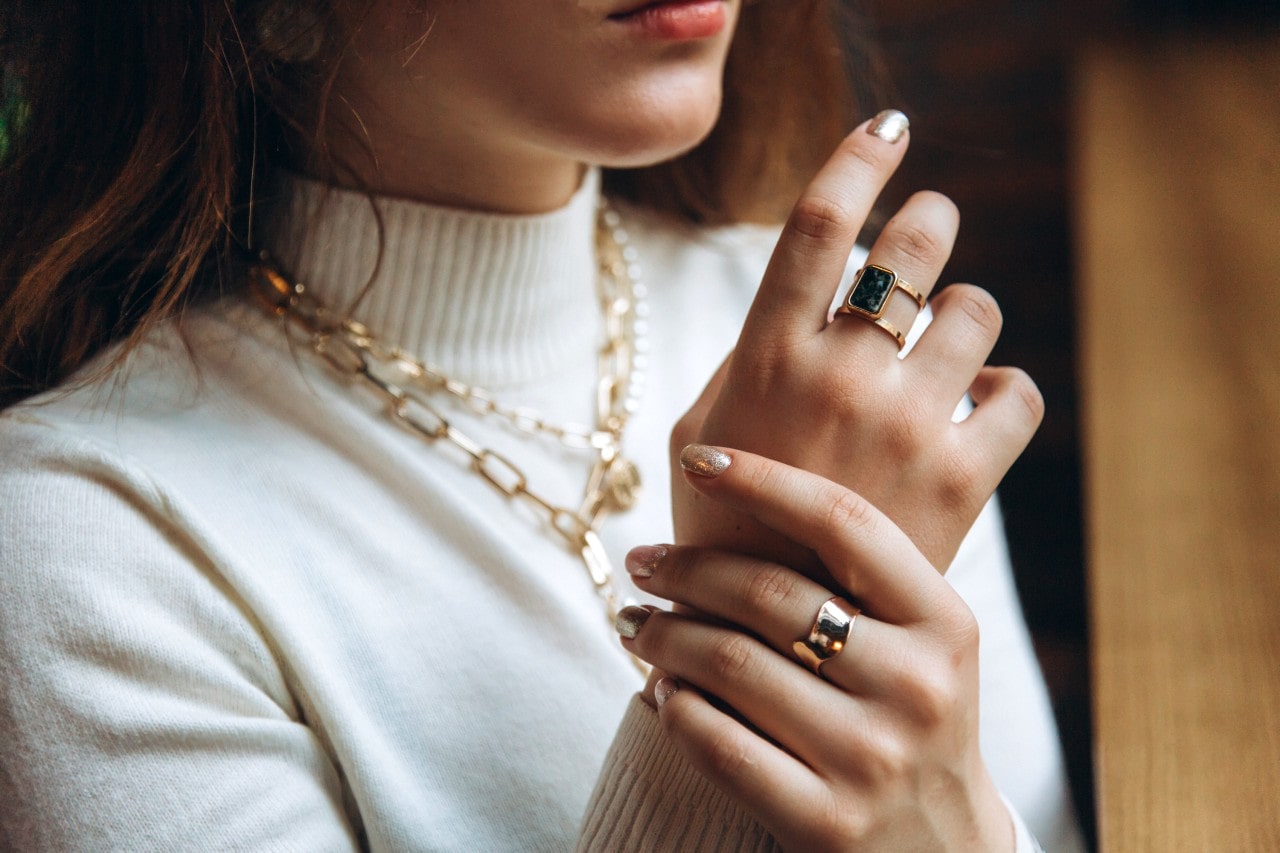 A woman in white turtleneck showcases yellow gold bulky chain necklaces and bold fashion rings