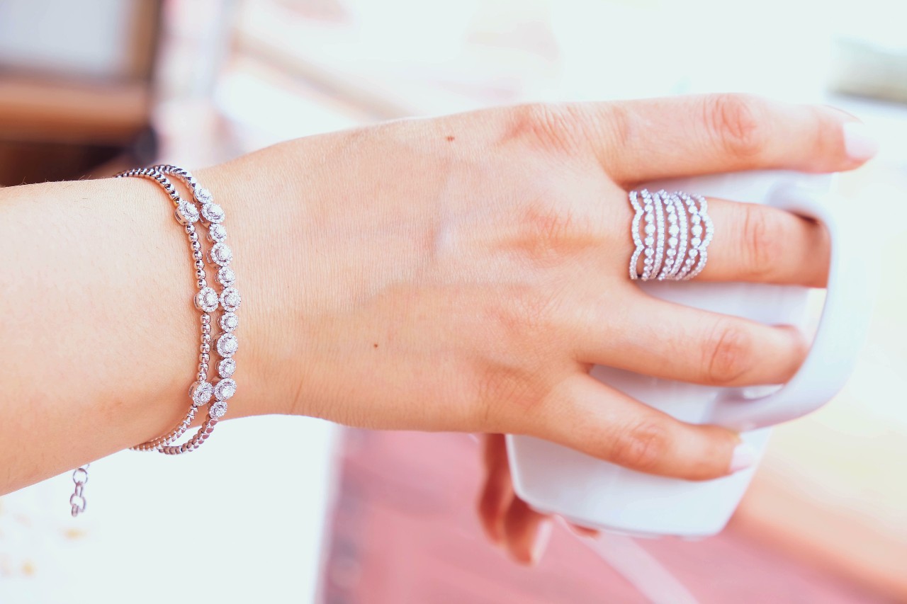 A hand adorned with a sparkling diamond bracelet and a multi-row diamond ring gracefully holds a white cup