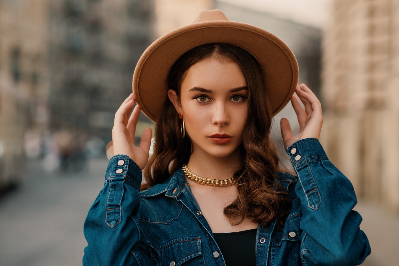 A young woman in a denim shirt and brown hat wearing a chunky gold chain necklace and hoop earrings.
