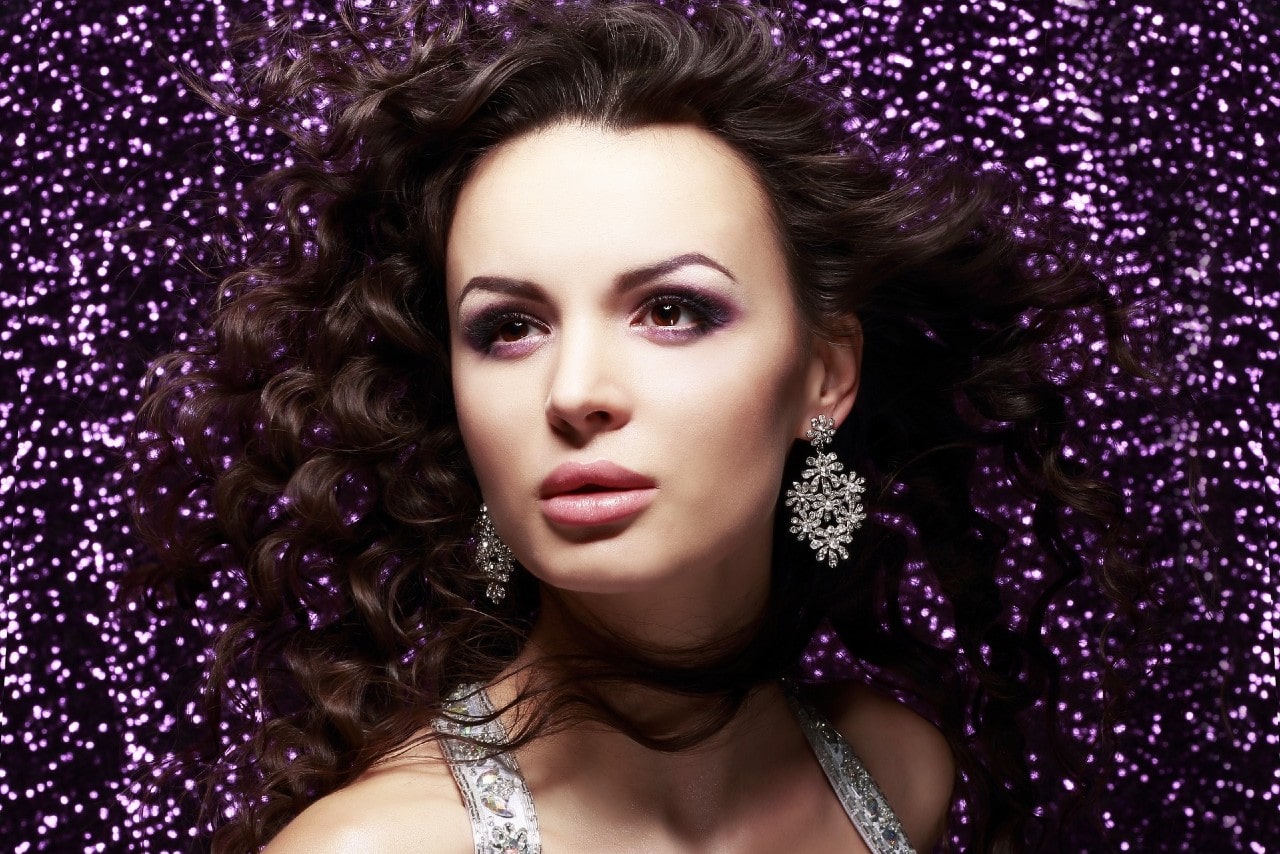 A portrait of a woman with voluminous curly hair, wearing ornate, sparkling chandelier earrings against a shimmering purple background. A portrait of a woman with voluminous curly hair, wearing ornate, sparkling chandelier earrings against a shimmering purple background.