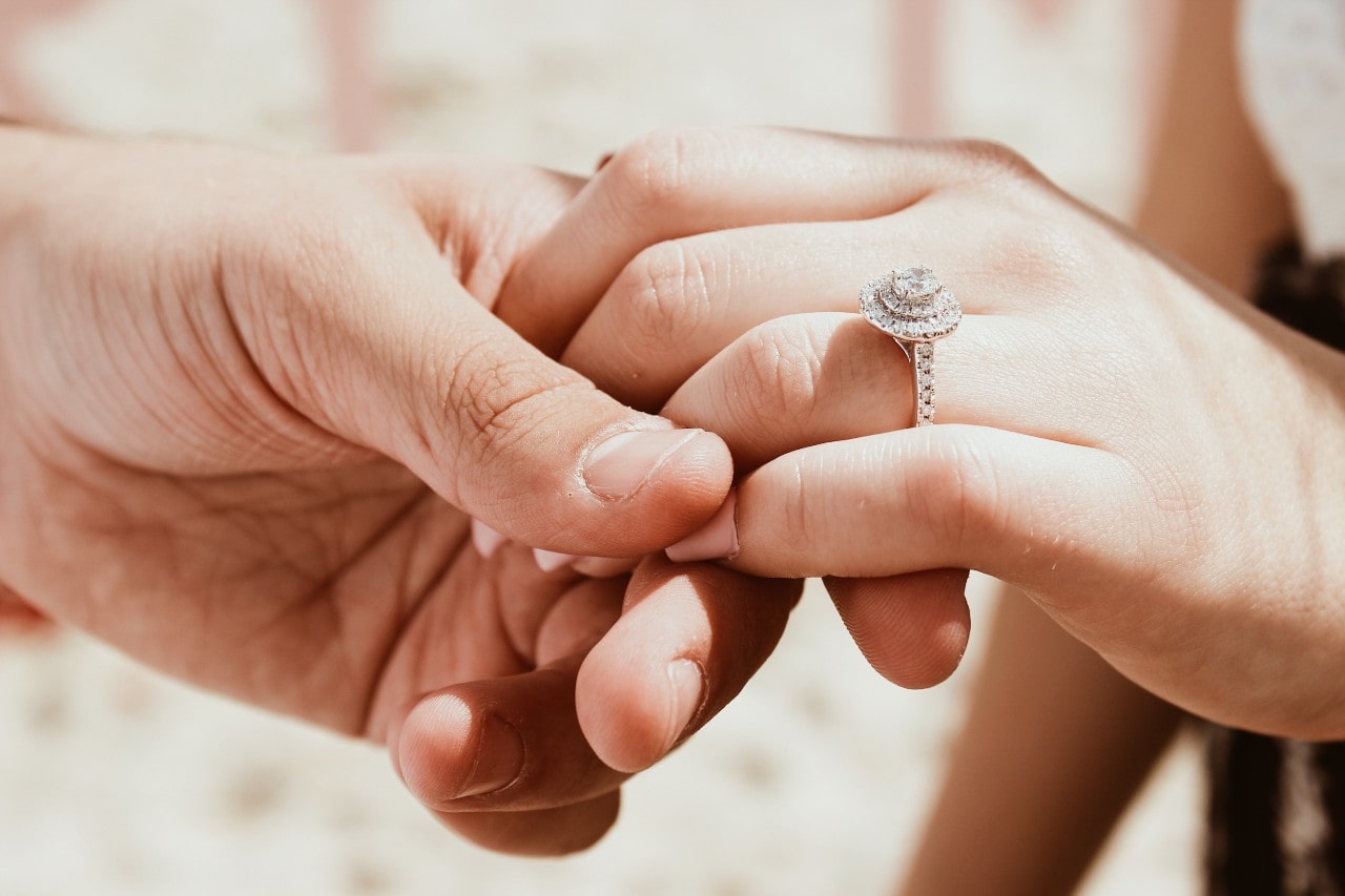A close-up of two hands clasped together, with a sparkling halo-style diamond engagement ring prominently displayed on a bride’s finger. A close-up of two hands clasped together, with a sparkling halo-style diamond engagement ring prominently displayed on a bride’s finger.