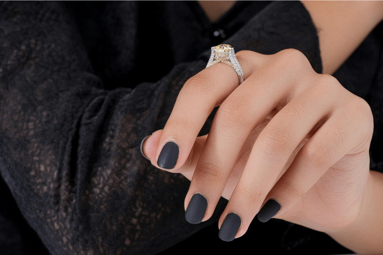 A close-up of a woman’s hand with a matte black manicure wearing an ornate side stone engagement ring.