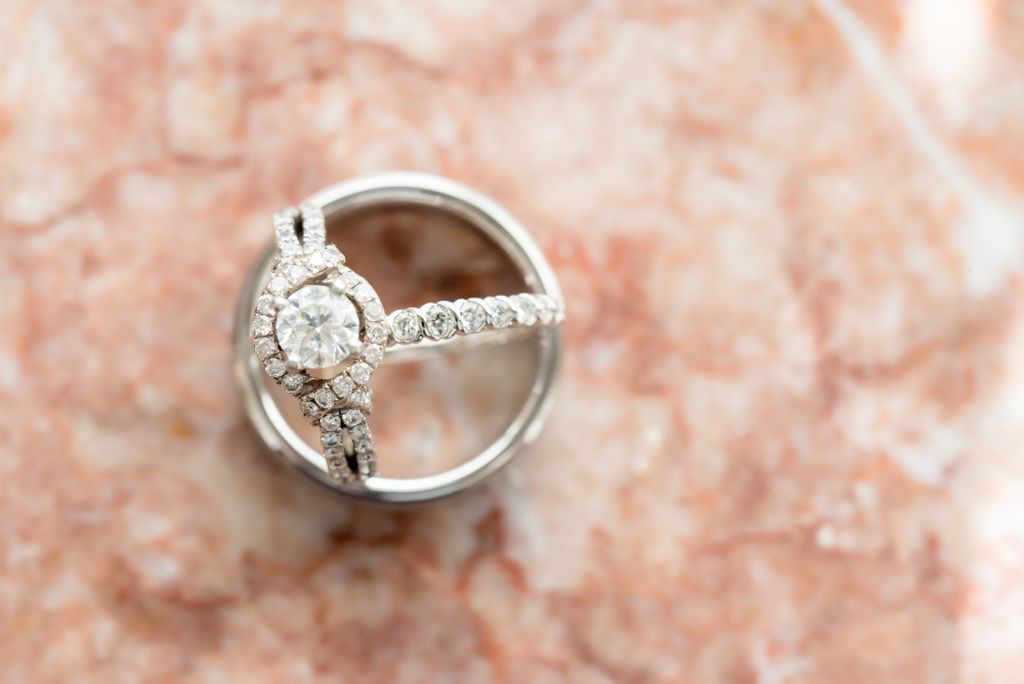 A close-up of a side stone halo engagement ring and wedding band pair displayed inside a men’s wedding band on a marble backdrop.