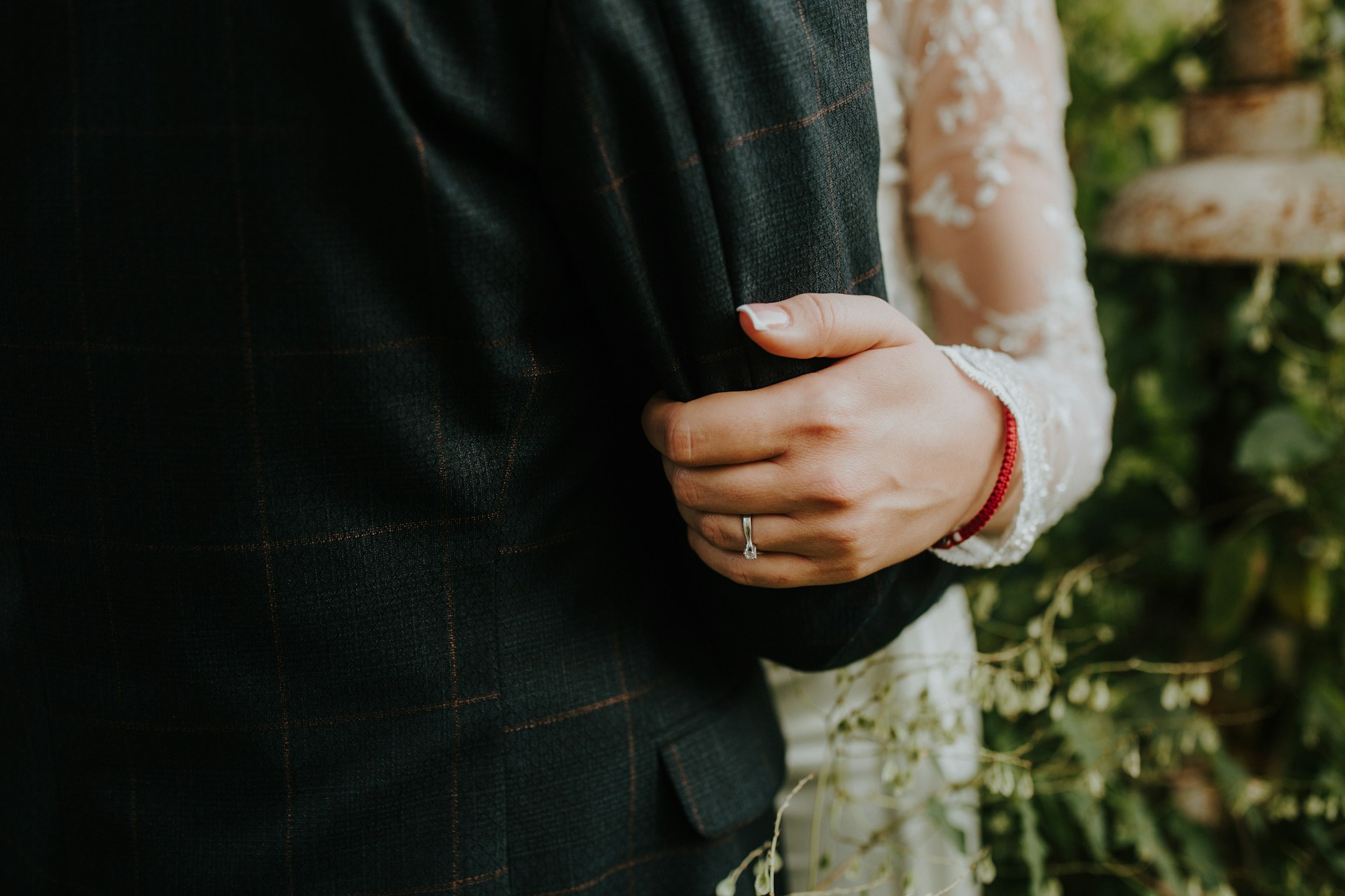 A close up of a bride in a lace wedding gown holding a groom by the elbow, showcasing a diamond wedding band.