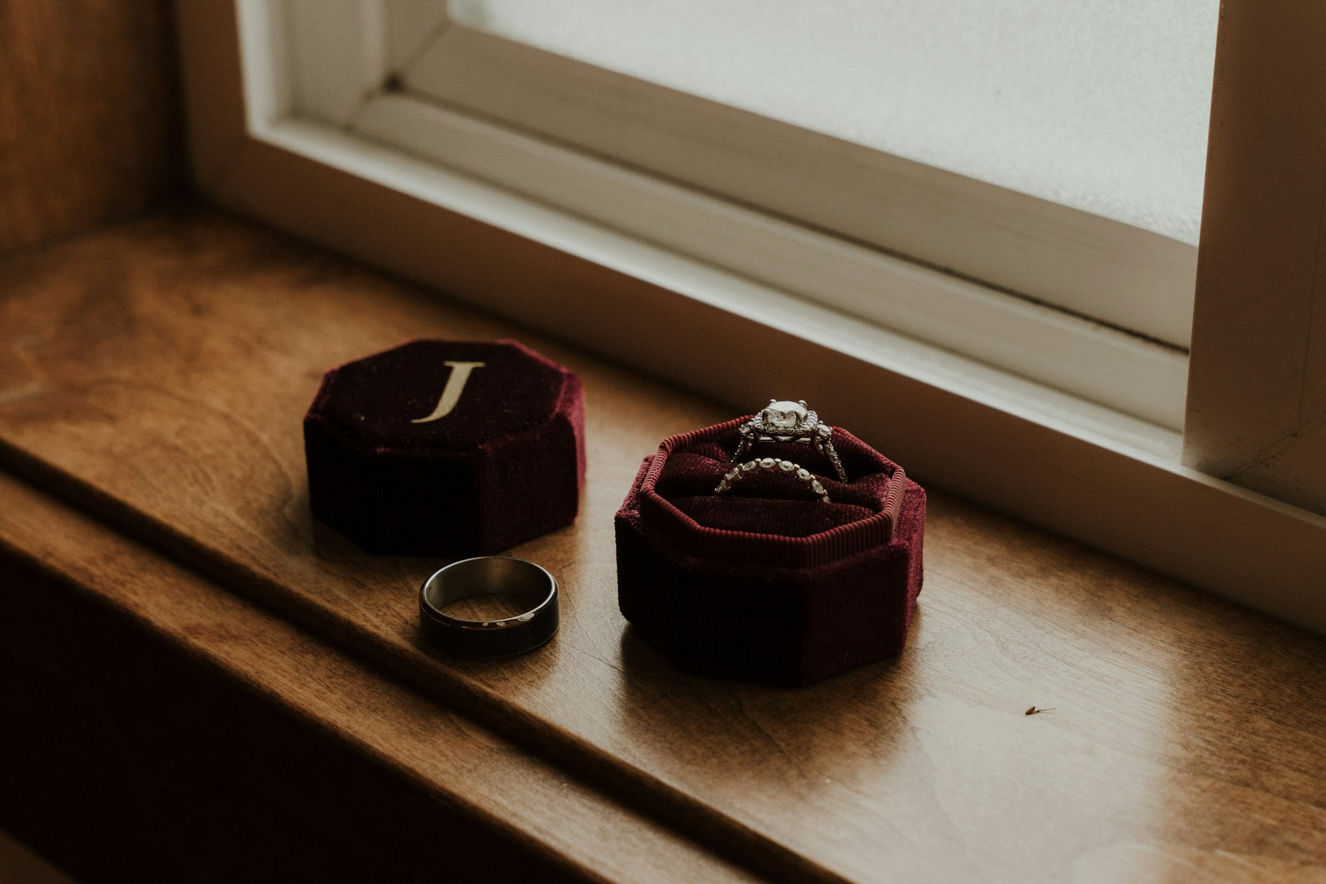 A velvet ring box sitting on a windowsill containing an engagement ring and matching band, and a men’s wedding band next to the box. A velvet ring box sitting on a windowsill containing an engagement ring and matching band, and a men’s wedding band next to the box.