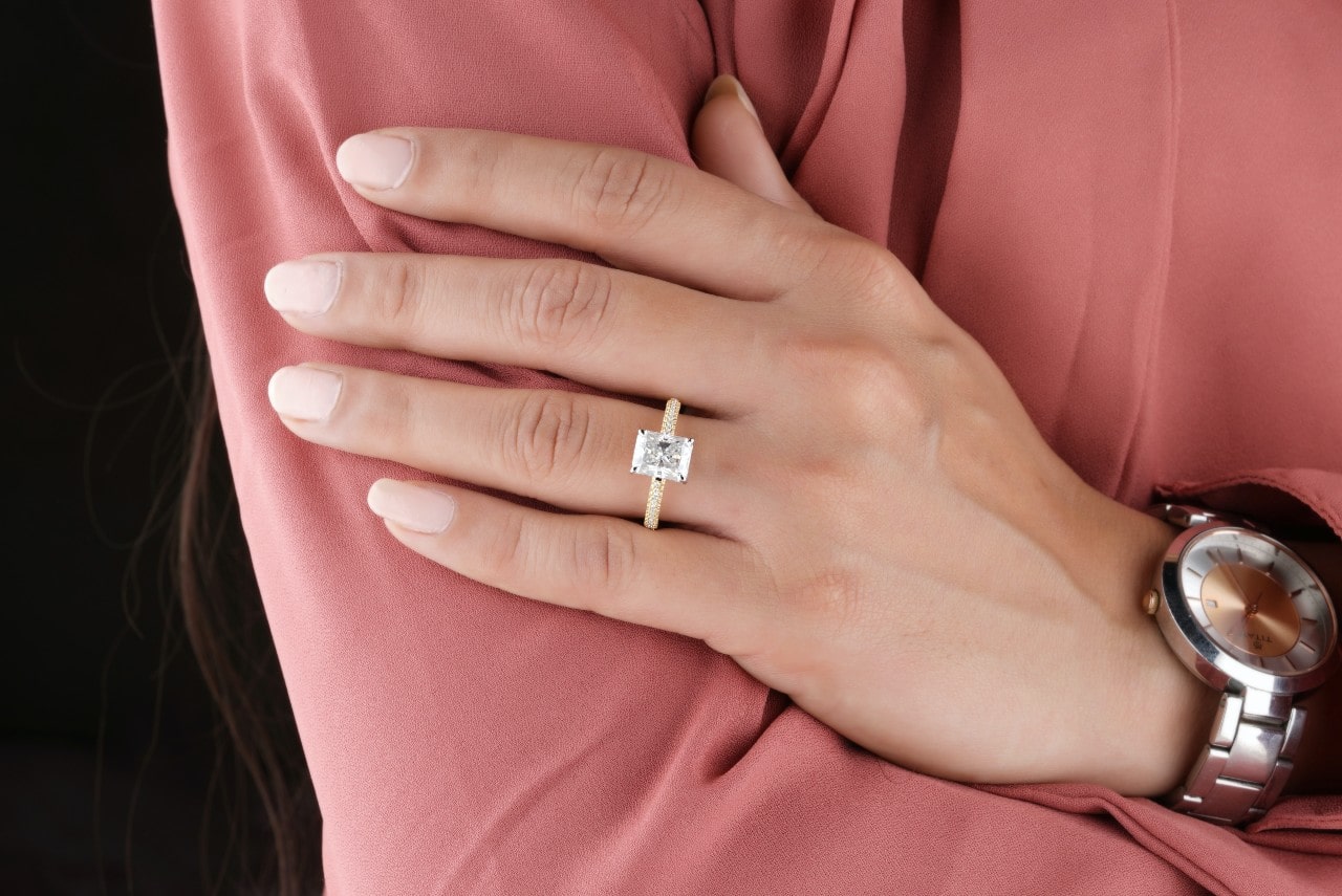 A woman dressed in pink crossing her arms, displaying a radiant-cut diamond engagement ring with a gold band. A woman dressed in pink crossing her arms, displaying a radiant-cut diamond engagement ring with a gold band.