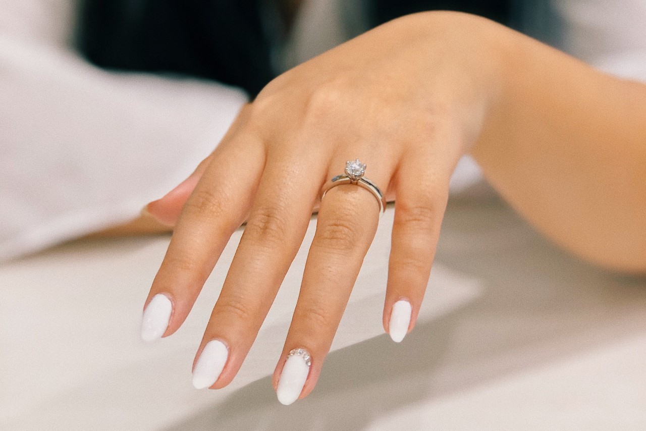 A hand with white nails showcasing a classic solitaire engagement ring on a silver band. A hand with white nails showcasing a classic solitaire engagement ring on a silver band.