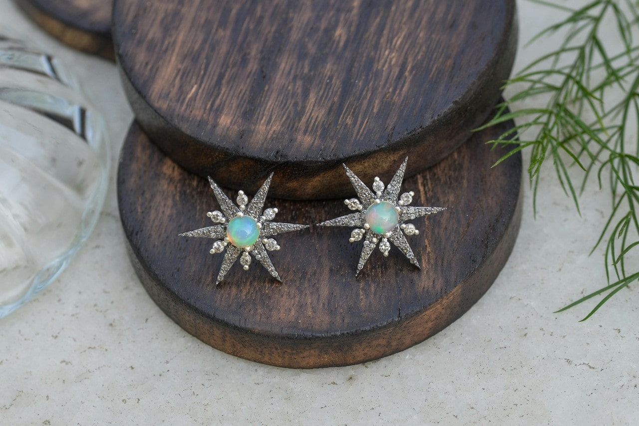A close-up of a pair of star-shaped stud earrings with opal center stones on a round piece of wood