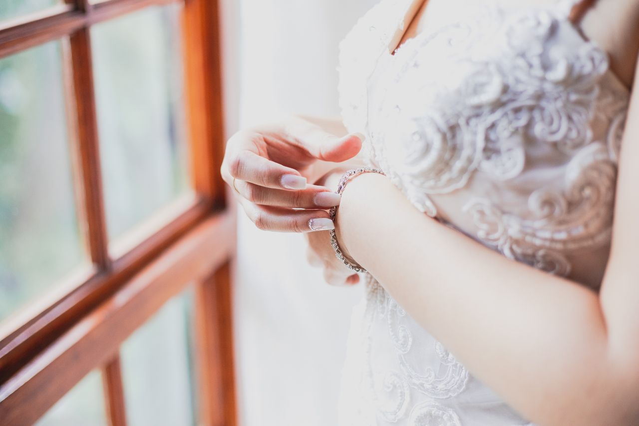 A bride in a detailed white lace gown stands by a window, adjusting a pearl bracelet.