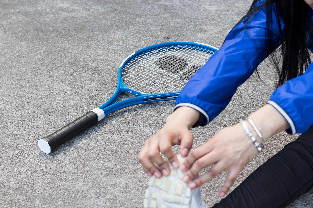 A person stretching on a court with a blue tennis racket lying on the ground. Their wrist features a silver tennis bracelet paired with a slim silver bangle.